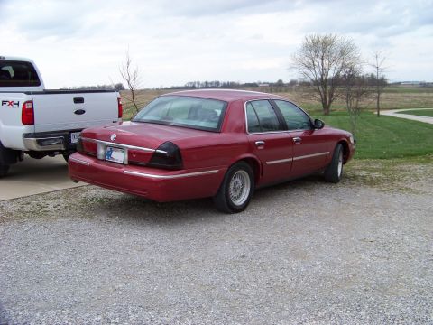 1999 Mercury Grand Marquis - A red sedan is parked on a gravel lot next to a white pickup truck under a cloudy sky.