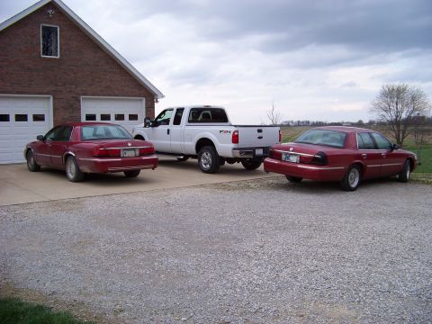1999 Mercury Grand Marquis - Two red 1999 Mercury Grand Marquis sedans and a white Ford pickup truck are parked in a driveway next to a garage.
