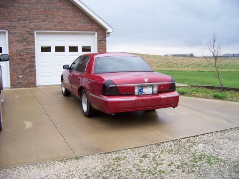 1999 Mercury Grand Marquis - Rear view of a red sedan parked in a driveway with a brick garage and rural background.