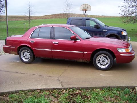 1999 Mercury Grand Marquis - A red sedan is parked on a concrete driveway next to a blue pickup truck, with a basketball hoop visible.