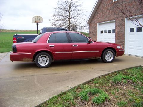 1999 Mercury Grand Marquis - A red sedan is parked on a concrete driveway next to a brick house with two garage doors.
