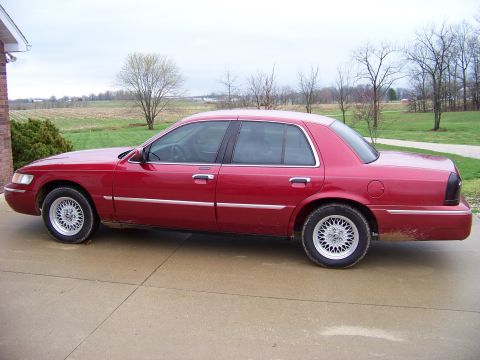 1999 Mercury Grand Marquis - Side view of a red Mercury Grand Marquis sedan. The car is parked on a concrete surface with a rural landscape behind it.