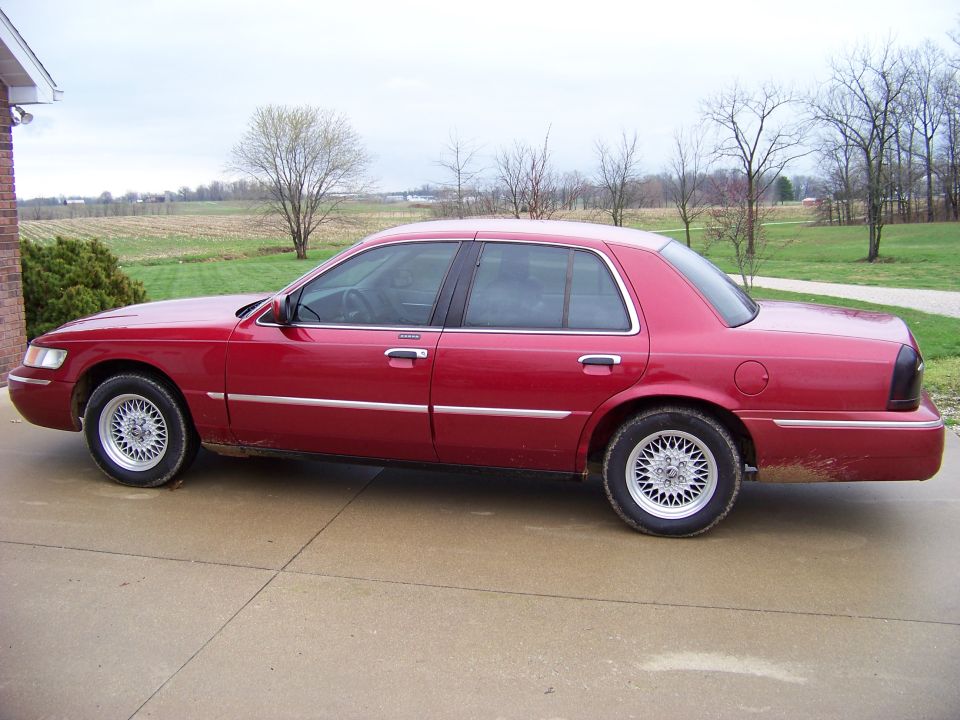 Red Mercury Grand Marquis sedan parked on concrete driveway, rural background