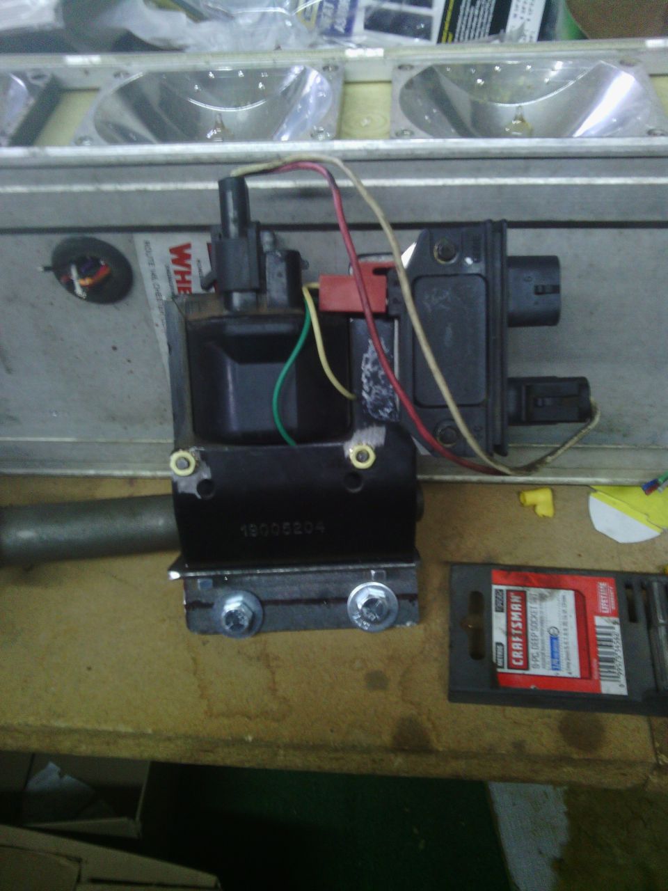 Ignition coil with wires attached, mounted on a metal bracket with screws. A Craftsman tool package is to the right