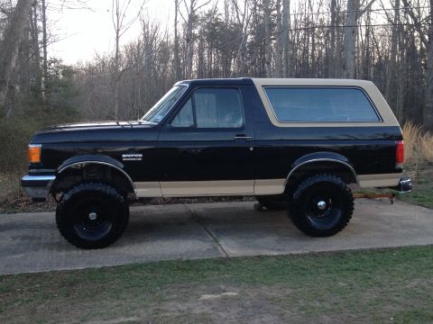 1991 Ford Bronco - A black and tan 1991 Ford Bronco is parked on a concrete driveway, its side profile clearly visible.