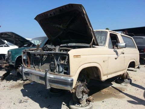 1995 Ford Bronco - This tan SUV, likely from the early 1980s, sits in a junkyard with its hood open and wheels removed.