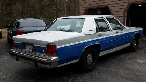1991 Ford LTD Crown Victoria - A two-tone blue and white Ford Crown Victoria police car is parked next to another dark-colored vehicle.