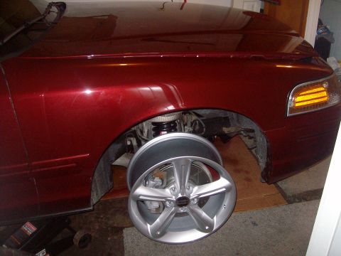 2004 Ford Crown Victoria - Dark red sedan raised on a jack in a garage with the front passenger wheel removed, exposing the coil spring, suspension, and brake assembl…