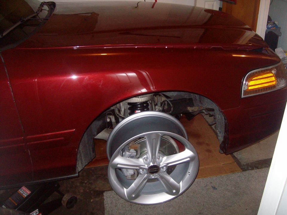 Dark red sedan on jack with front wheel off and silver alloy wheel leaning nearby