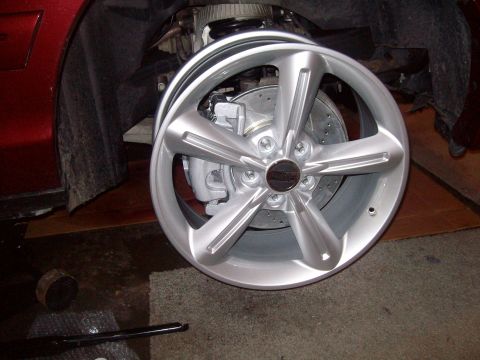 2004 Ford Crown Victoria - A silver five-spoke alloy wheel with a black center cap leans bare against the jacked-up front corner of a dark red car in a garage, exposi…