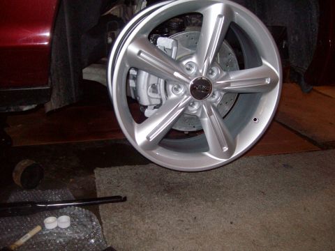 2004 Ford Crown Victoria - Bare silver five-spoke alloy wheel with a chrome center cap propped against the front of a dark red car in a garage, with the drilled-and-s…