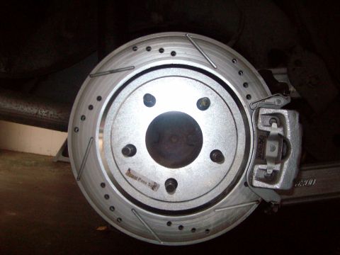 2004 Ford Crown Victoria - Head-on close-up of a newly fitted cross-drilled and slotted brake rotor labeled "Front Pass Side" over exposed wheel studs, with a bare ca…