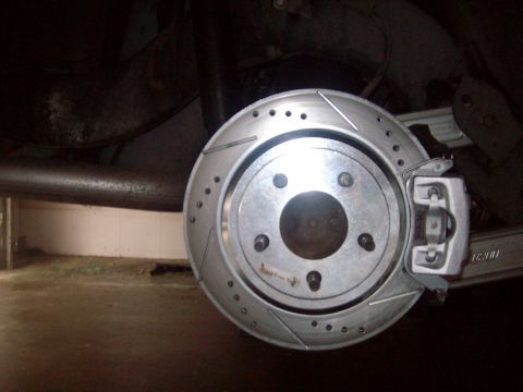 2004 Ford Crown Victoria - Close-up of a newly installed cross-drilled and slotted brake rotor with a bare-metal caliper on a rear hub, wheel removed, photographed fr…