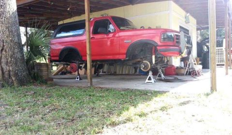 1995 Ford Bronco - A red Ford Bronco is elevated on jack stands for maintenance work beneath a carport.