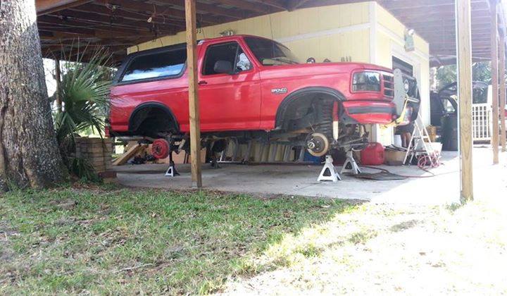 Red Ford Bronco on jack stands, undergoing repairs under a covered carport
