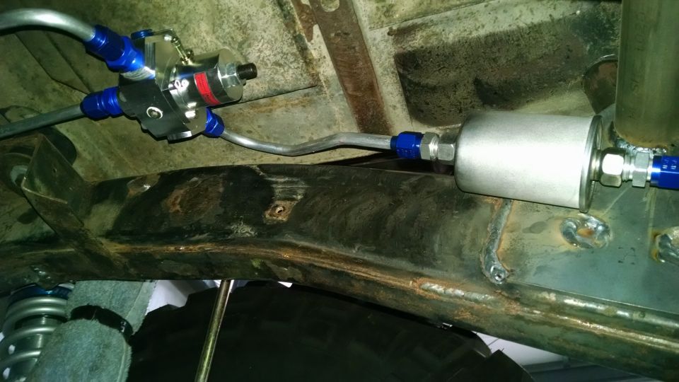 Underside view of a fuel filter and regulator assembly with blue fittings attached to rusty frame