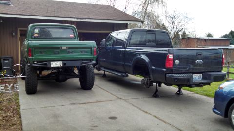 1967 Ford F-250 - Two Ford trucks are parked in a driveway, one a classic green model and the other a newer dark blue model missing its wheels.