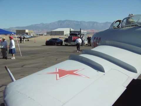 1995 Peterbilt 379 - A fighter jet's wing is in the foreground, with a semi-truck and a car visible on the tarmac in the background.