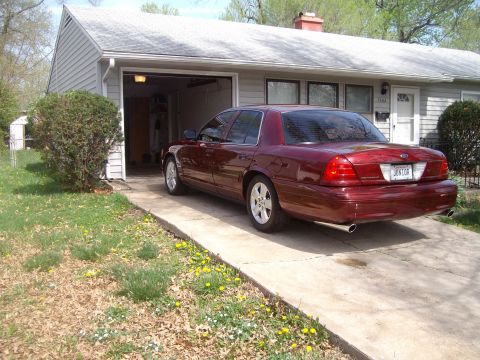 2004 Ford Crown Victoria - Three-quarter rear view of a dark red Ford Crown Victoria sedan parked on a concrete driveway at the mouth of an open attached garage, wear…