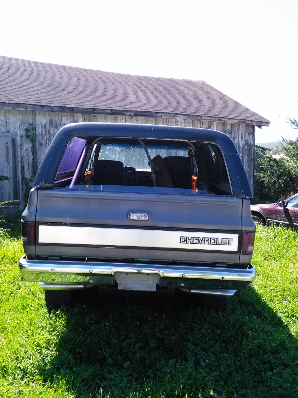 Rear view of a dark blue Chevrolet K5 Blazer parked on grass in front of a barn. Another car is visible to the right