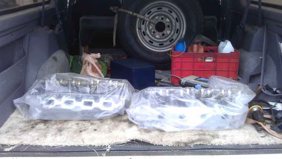 Two engine camshafts and exhaust manifolds wrapped in plastic, stored in the cargo area of a vehicle with a spare tire and red crate