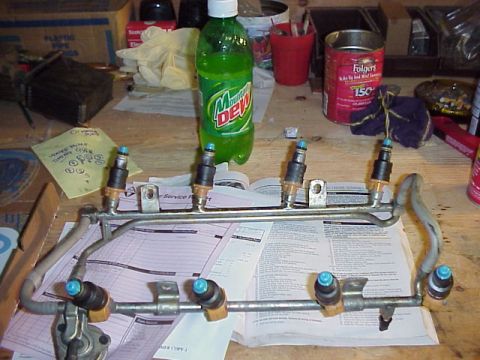 1989 Ford Bronco - A fuel injector rail sits on a table covered with papers, next to a bottle of Mountain Dew and a can of coffee.