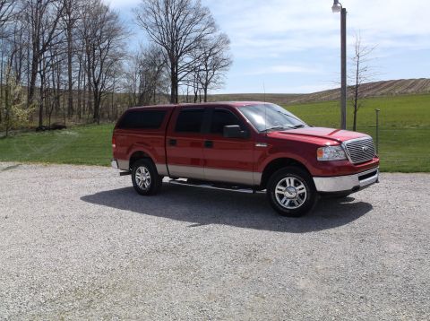 2008 Ford F-150 - A red pickup truck with a camper shell is parked on a gravel lot with trees and a field in the background.