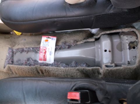 1999 Mercury Grand Marquis - Car interior floor with carpet removed, revealing padding and a console. A package of eye drops is on the floor.