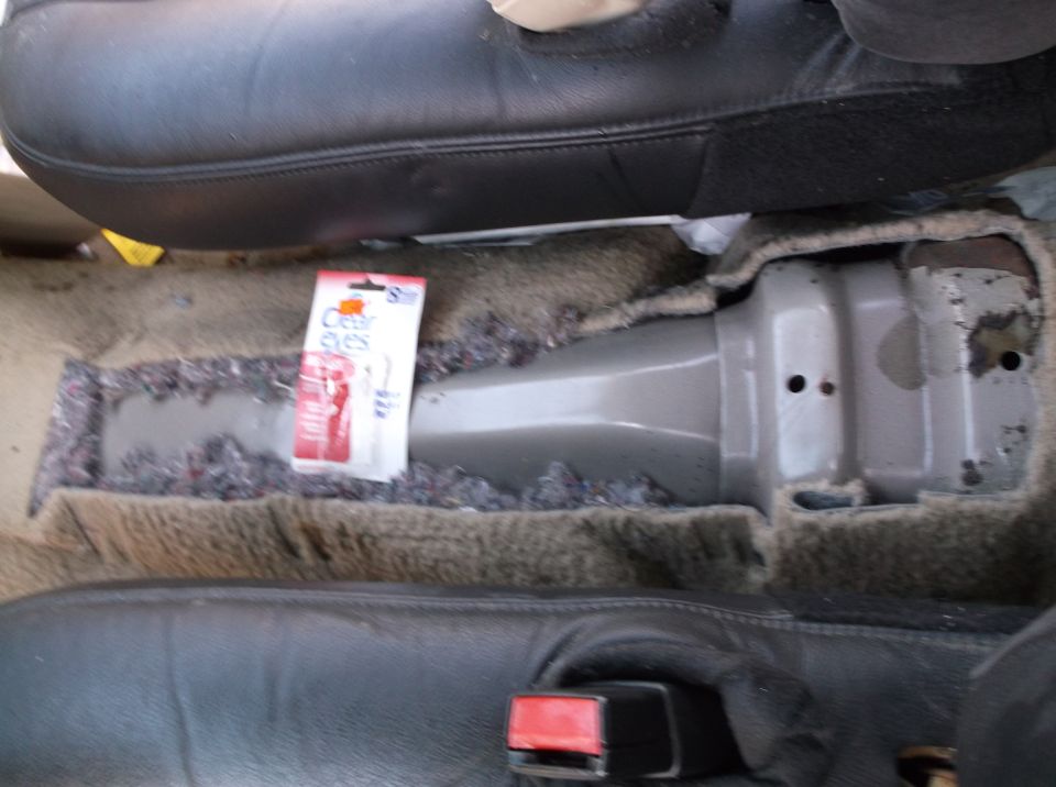 Car floor showing exposed carpet padding and a package of Clear Eyes eye drops next to a gray metal console. Seatbelt buckle visible