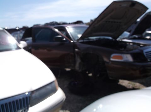 1999 Mercury Grand Marquis - A 1999 Mercury Grand Marquis sits in a junkyard with its hood ajar, alongside other vehicles.