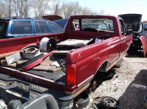 1999 Mercury Grand Marquis - A red pickup truck sits in a junkyard with various parts in its bed and other vehicles visible.