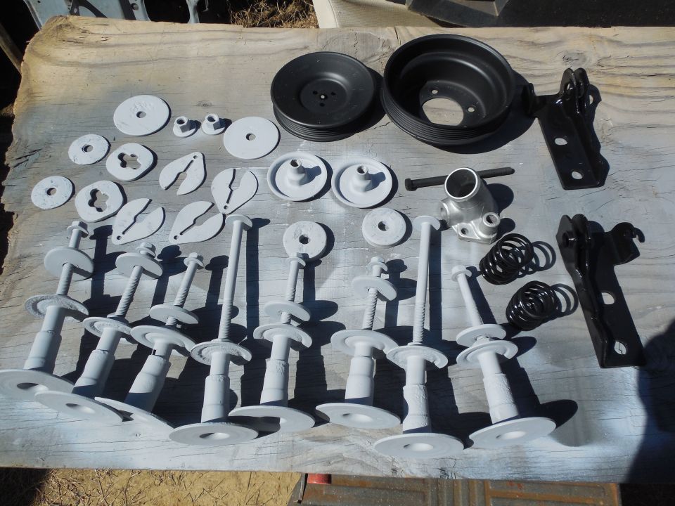 Gray-painted bolts, plates, pulleys, springs and brackets arranged on a wood board outdoors.
