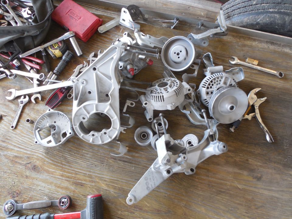 Disassembled engine brackets, alternators and pulleys on wood with scattered hand tools.