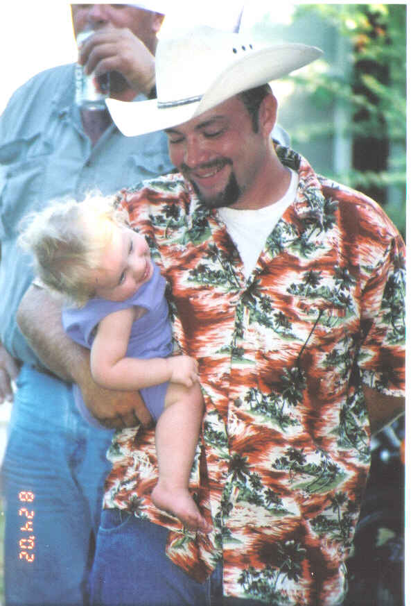 Man in a Hawaiian shirt holding a baby, with another man in the background