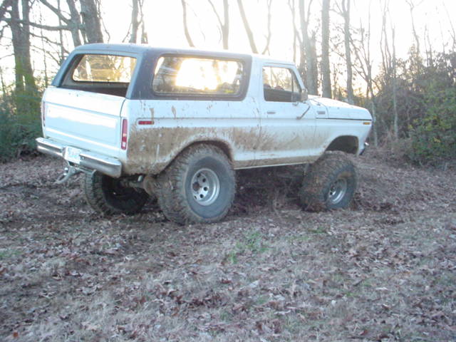 White 1978 Ford Bronco with mud splatters, parked on a dirt path with trees in the background