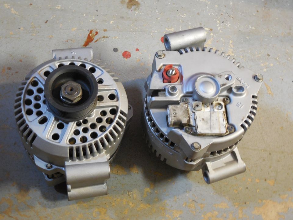 Two silver car alternators on concrete, one showing pulley, one showing rear terminals