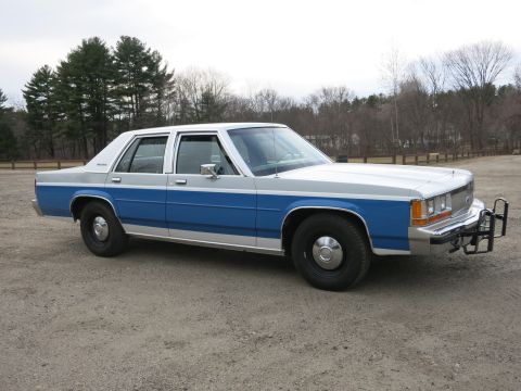1991 Ford LTD Crown Victoria - A two-tone blue and silver Ford Crown Victoria sedan is parked on a gravel lot with trees in the background.