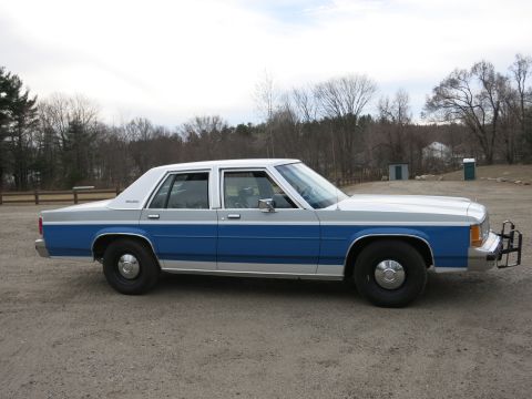 1991 Ford LTD Crown Victoria - A two-tone blue and silver sedan with a push bumper is parked on a gravel lot.
