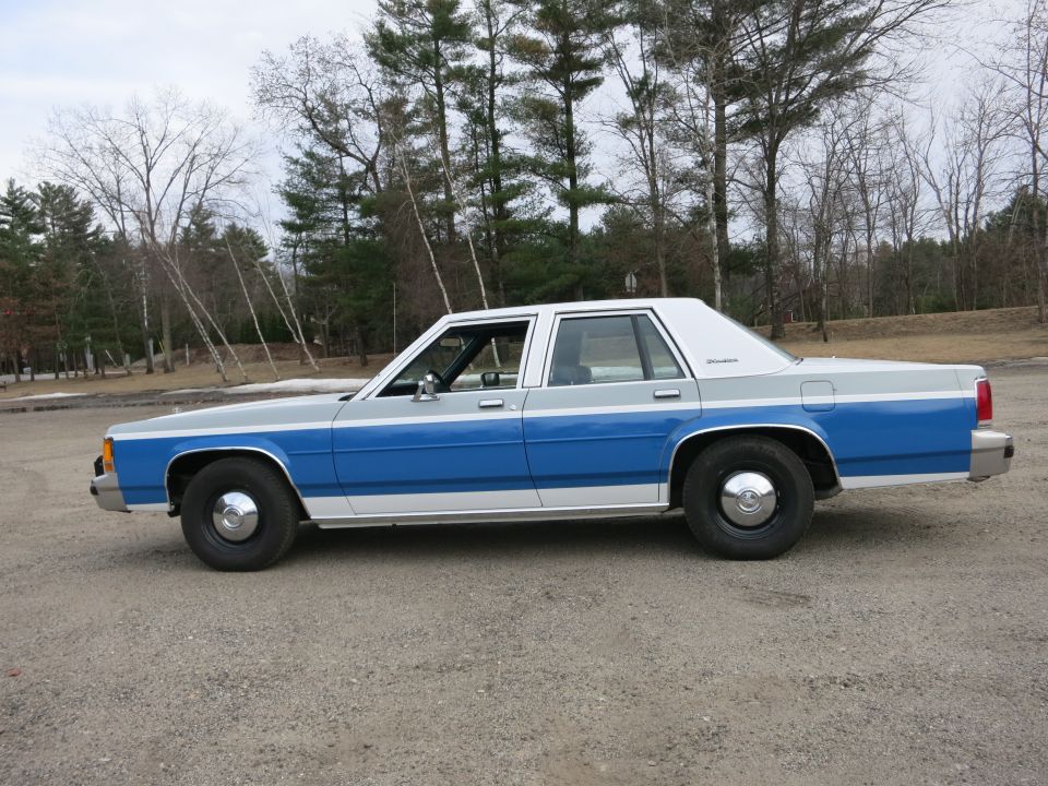Side view of a two-tone blue and gray sedan with chrome hubcaps parked on a gravel lot