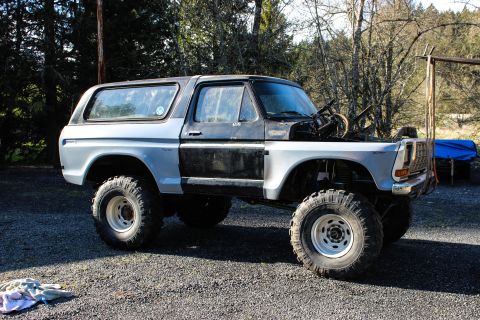 1979 Ford Bronco - Driver-side profile of a stripped, lifted full-size two-door Bronco-style SUV parked on gravel in front of trees, with a white/silver lower…