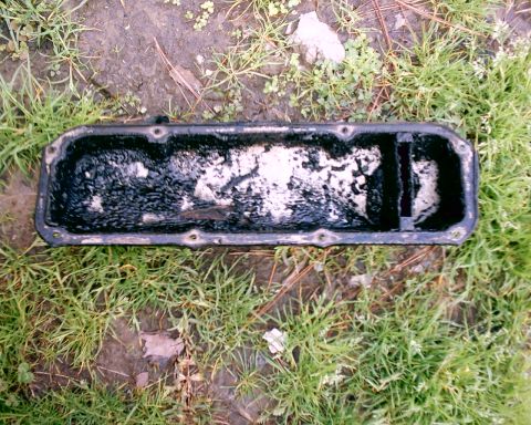 1980 Ford Bronco - A used valve cover with significant internal buildup is shown resting on a patch of grass and dirt.