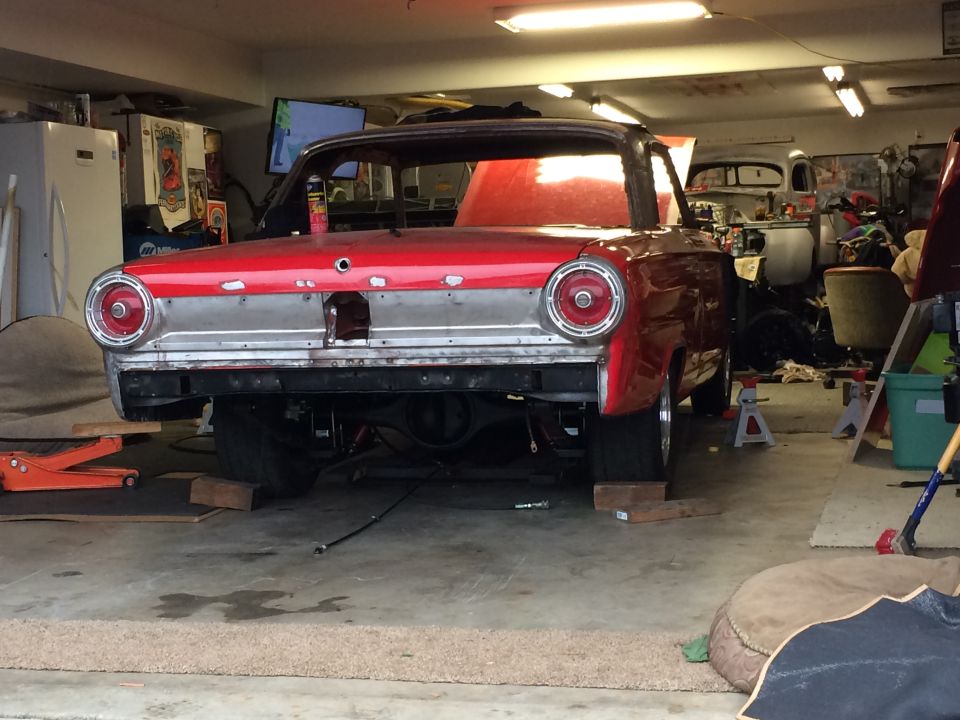 1964 Ford Fairlane coupe in garage, rear view, undergoing restoration. Another vehicle is visible in the background