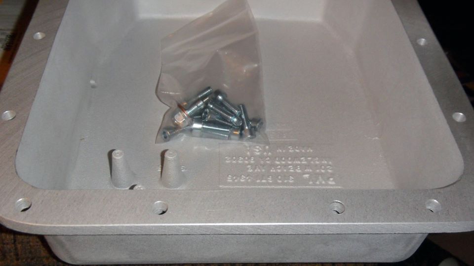 Aluminum transmission pan with a bag of bolts inside. Text is visible on the pan