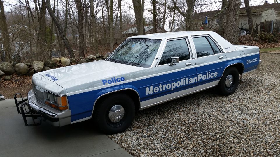White and blue police car, 1991 Ford Crown Victoria LTD, parked on gravel. "Metropolitan Police" text visible