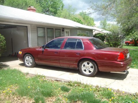 2004 Ford Crown Victoria - A red sedan is parked in a driveway in front of a house. The vehicle has tinted windows and alloy wheels.