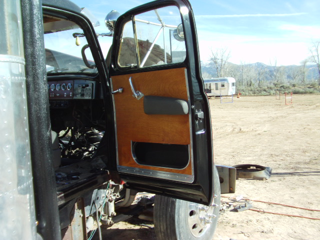 Open driver's door of a vintage truck showing wood-paneled door, dashboard gauges, and exposed engine components