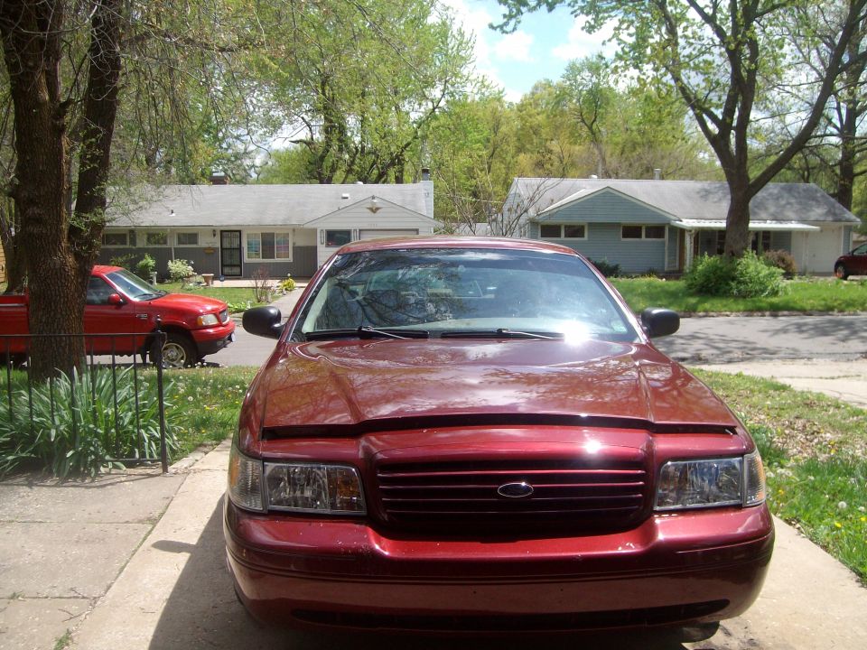 Red Ford Crown Victoria front view, hood slightly ajar. A red pickup truck is parked in the background