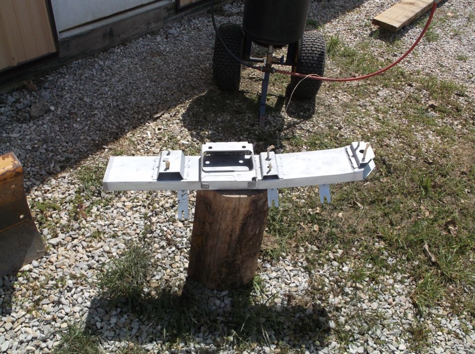 White painted car bumper assembly on a log, with a sandblaster in the background