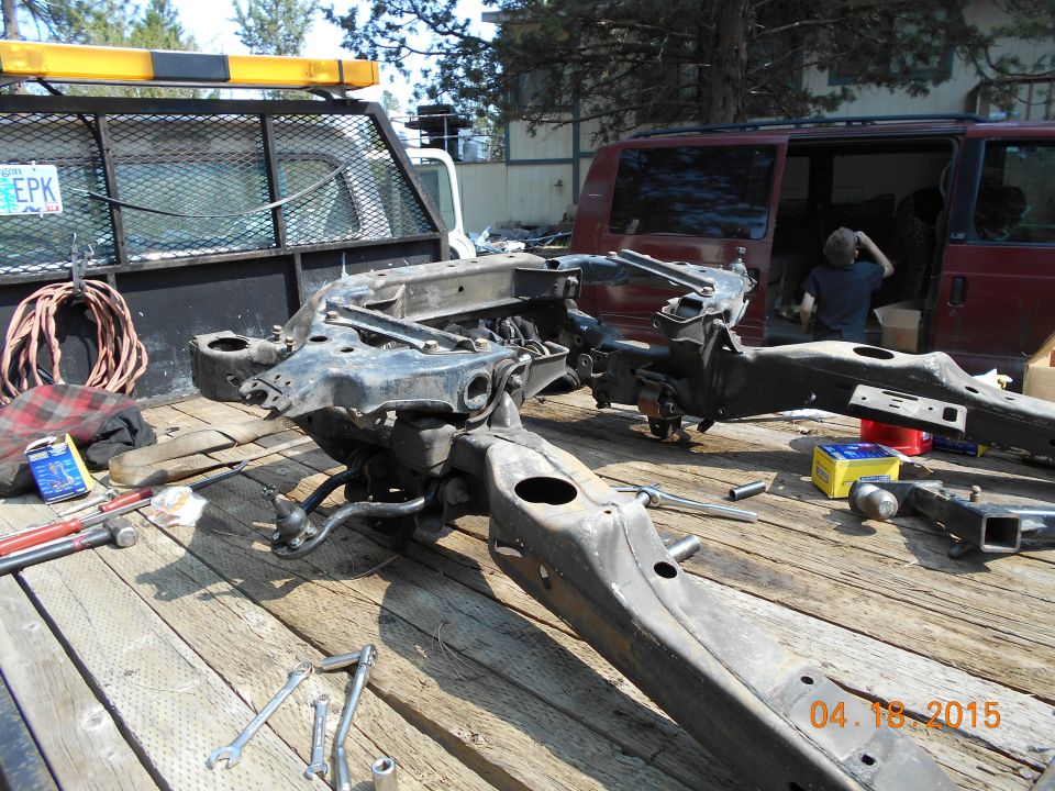 Vehicle frame and suspension components on a wooden trailer bed, with a maroon van and a truck in the background