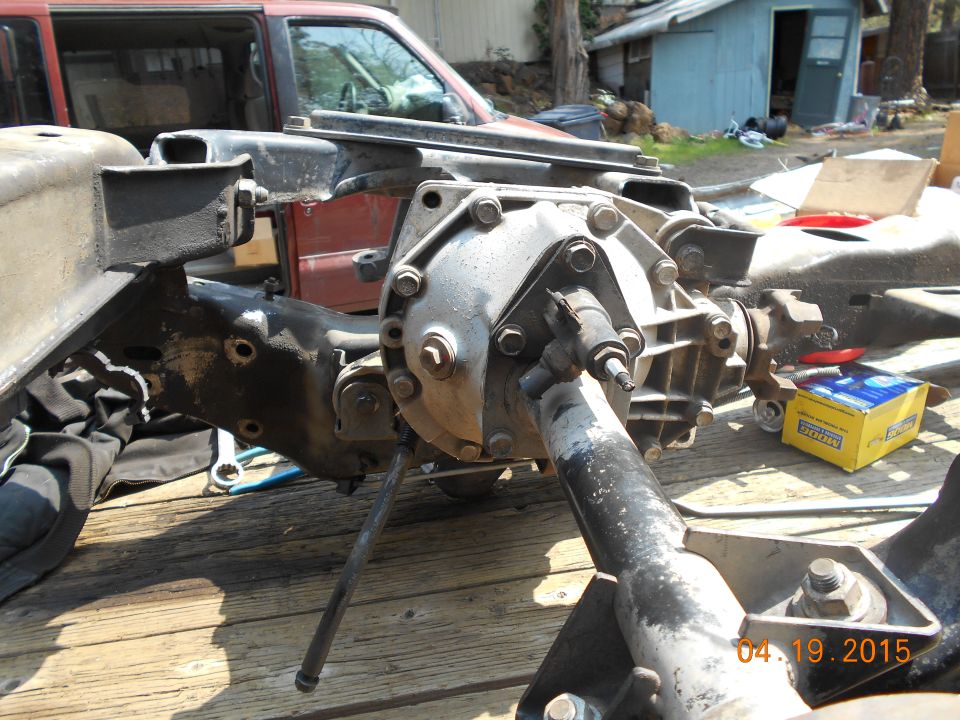 Differential and suspension components on wooden planks, with a red vehicle in the background